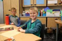 Two young boys sitting at a table in a classroom, engaged in an activity. One boy is writing on paper while the other looks