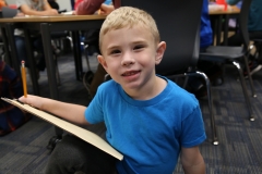Boy in blue shirt sitting at desk holding clipboard.