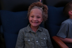 Young girl sitting in movie theater, smiling at camera, wearing green jacket and black pants.