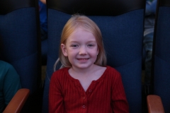 Little girl sitting in movie theater chair, smiling at camera during pen pal meet up day.