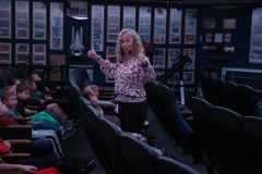 Woman standing in front of a group of children in an auditorium, gesturing with her hands.