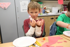 Two children at a table, engaged in an activity, with a teacher nearby observing. They are surrounded by paper materials and