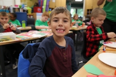 Young boy sitting at a table in a classroom, smiling at the camera, wearing a blue jacket and looking towards his pen pal.