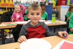 Young boy sitting at a table in a classroom, holding a plate of food, looking at the camera.