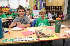 Two children sitting at a desk in a classroom, smiling and posing for a picture, surrounded by school supplies and crafts.