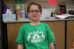 Boy in green shirt sitting at desk with sign that says "Sampson berry" on front of shirt.