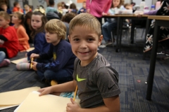 Boy sitting at table in classroom, smiling and holding pencil over paper.