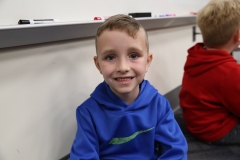 Young boy in blue jacket sitting at table smiling at camera.
