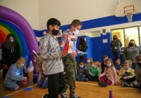 Two children in camouflage shirts standing at front of room, holding tablets, with colorful balloon arch behind them and