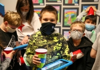 Children wearing masks, gathered around a table with cups and a tablet, possibly for an art project or learning activity.