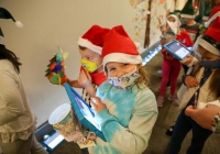 Children wearing santa hats, holding tablets, in a corridor.