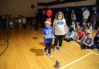 Two children in a gymnasium wearing masks, one child is holding a small cake with a candle on it and another child is